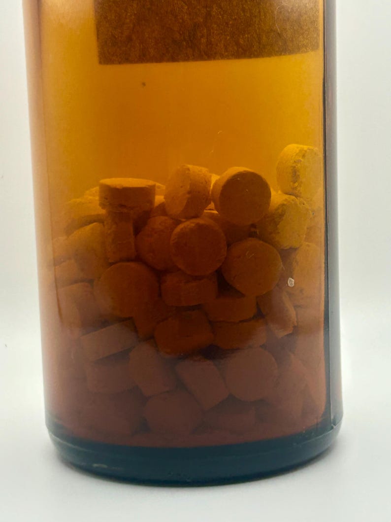 May include: A clear amber-colored bottle filled with numerous orange and white pills. The pills are small, round, and cylindrical. The bottle has a dark brown base and a label at the top.