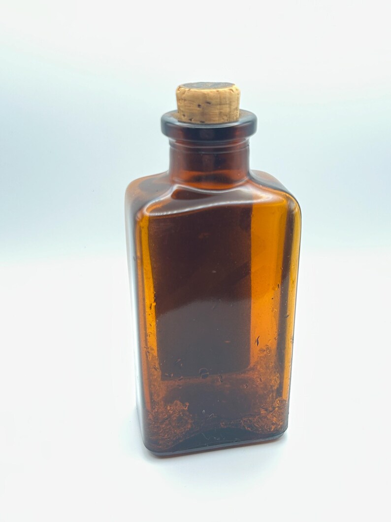 May include: A rectangular amber glass bottle with a cork stopper. The bottle has a square shape and a dark brown hue. The cork is light brown and fits snugly in the bottle's neck. The bottle is empty.