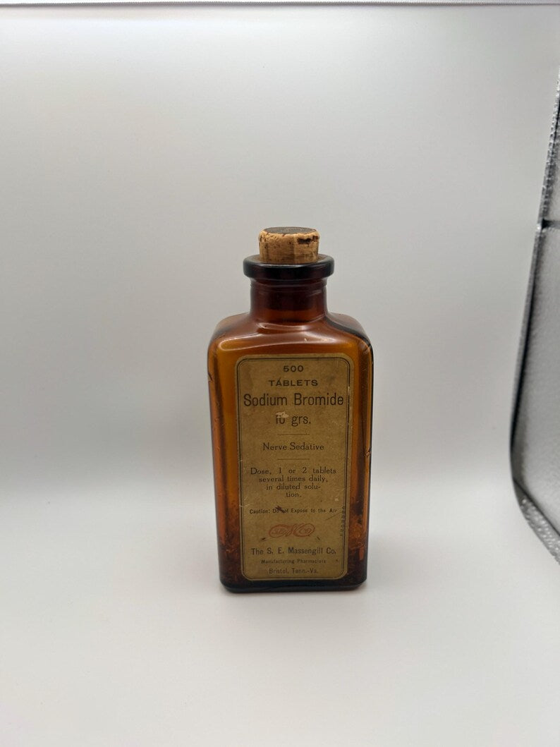 May include: An antique amber glass bottle with a cork stopper. The label reads "500 TABLETS Sodium Bromide 16 grs. Nerve Sedative." The bottle has a rectangular shape and is likely from the early 20th century. The label is aged and shows signs of wear.
