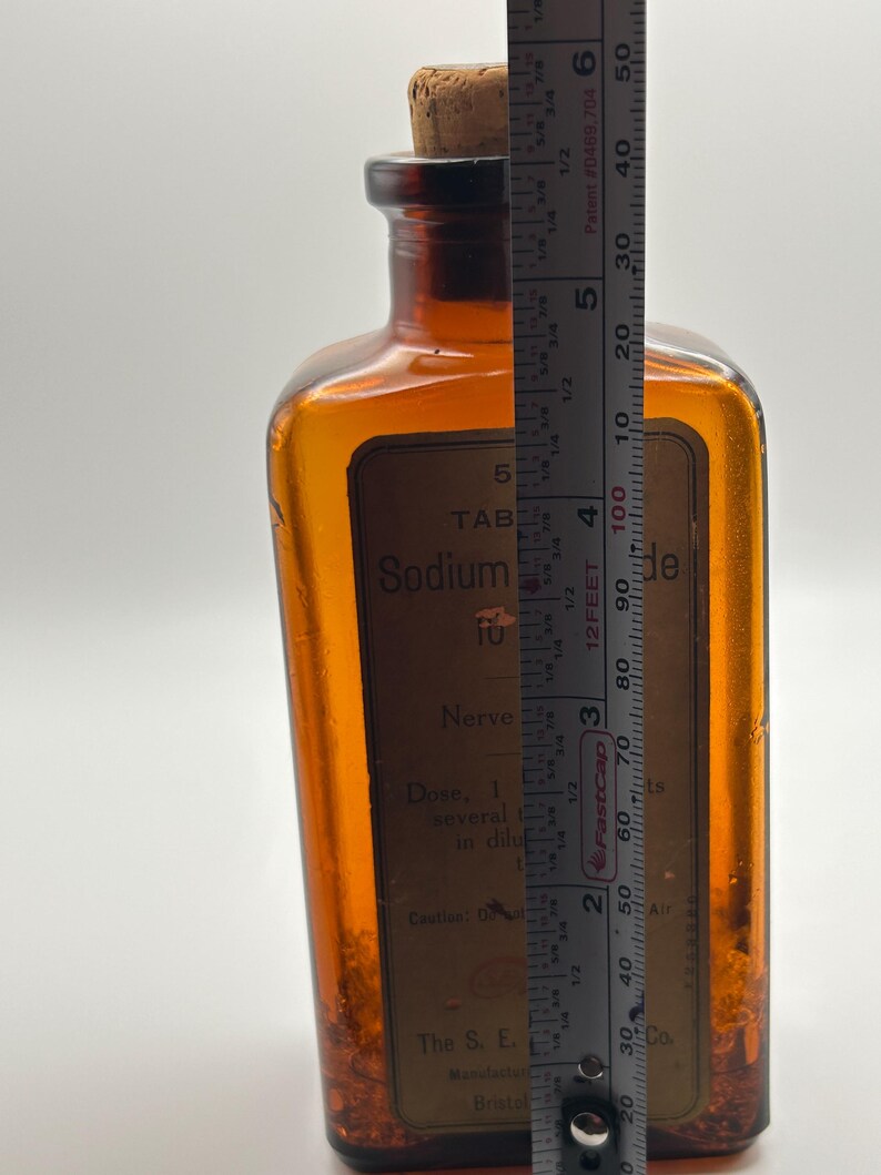 May include: An amber-colored glass apothecary bottle with a cork stopper. The bottle has a rectangular shape and a label with the text "Sodium" and "Nerve". The bottle is approximately 5 inches tall.