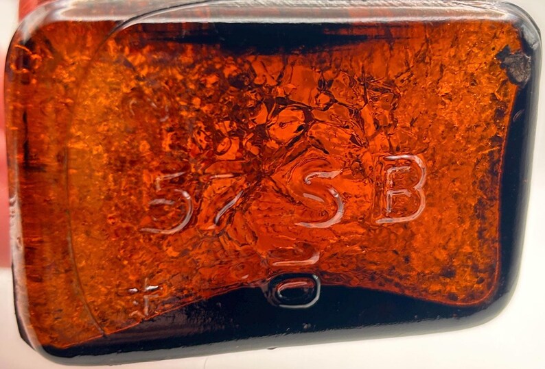 May include: Close-up of a rectangular amber-colored glass bottle. The bottle has embossed text: "57 SB 10". The glass has a textured surface and is filled with a dark liquid.