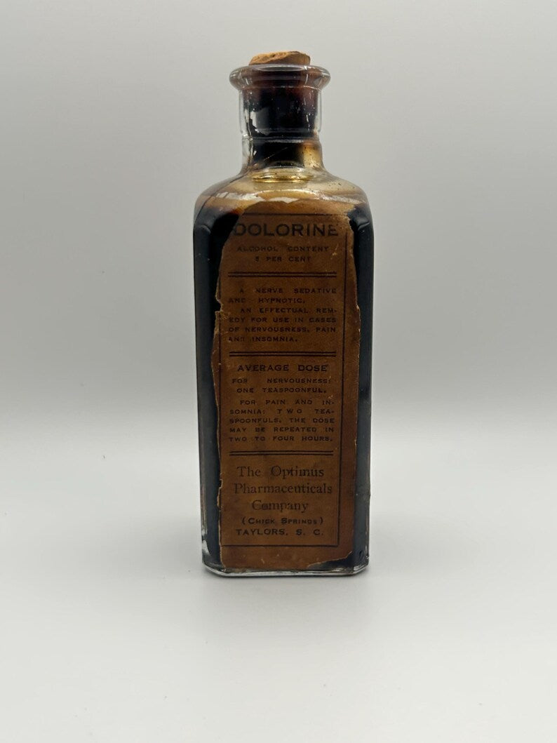 May include: A rectangular glass bottle filled with a dark liquid, topped with a cork stopper. A tan paper label reads DOLORINE, ALCOHOL CONTENT 5 PER CENT, A NERVE SEDATIVE AND HYPNOTIC, AVERAGE DOSE, The Optimus Pharmaceuticals Company, CHICK SPRINGS, TAYLORS, S. C.