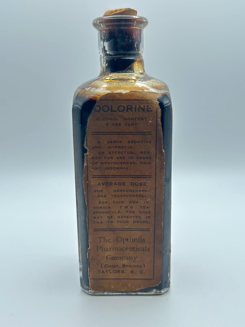 May include: A clear glass bottle filled with a dark liquid, sealed with a cork stopper. A tan paper label is adhered to the front, printed with the text DOLORINE, ALCOHOL CONTENT 5 PER CENT, A NERVE SEDATIVE AND HYPNOTIC, and The Optimus Pharmaceuticals Company.