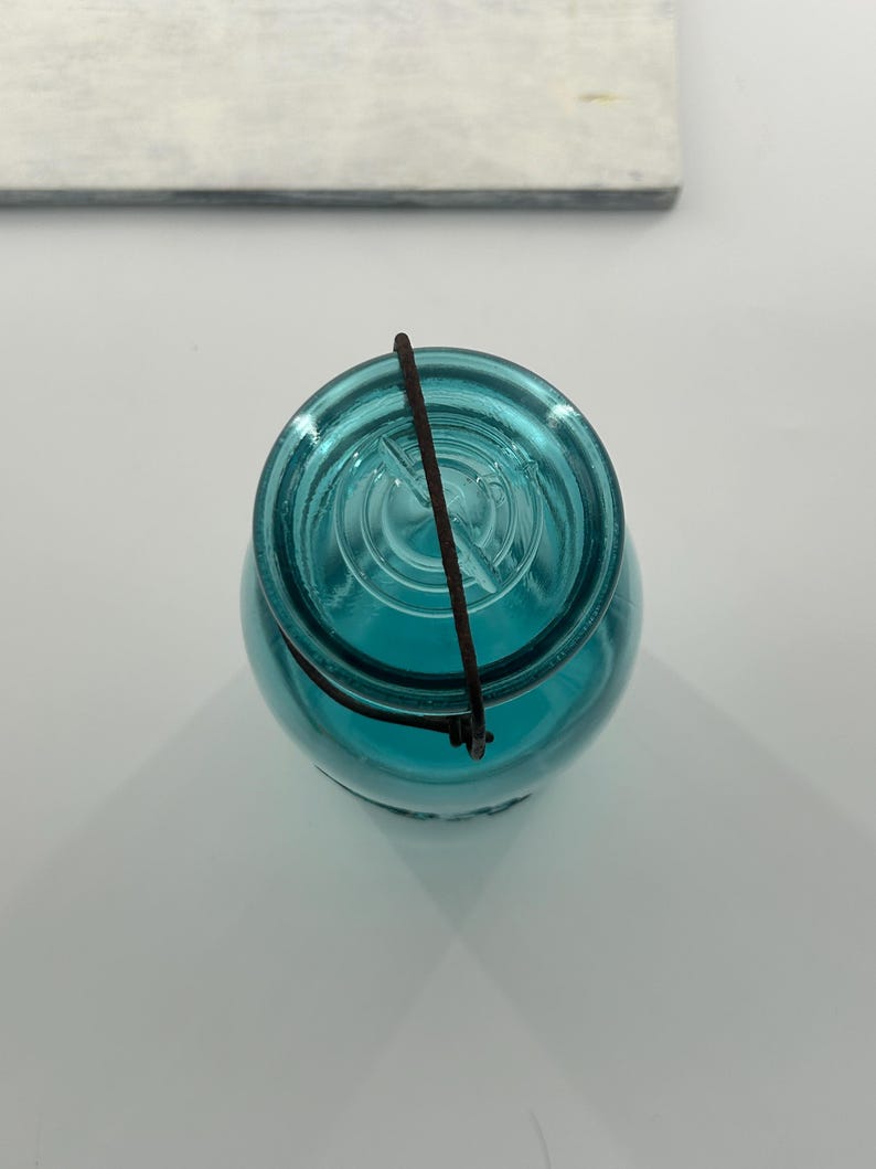 May include: A vintage, teal-colored glass jar with a wire closure. The jar has a rounded shape and a clear, textured surface. The wire closure is dark brown. The jar is likely used for canning or preserving food.