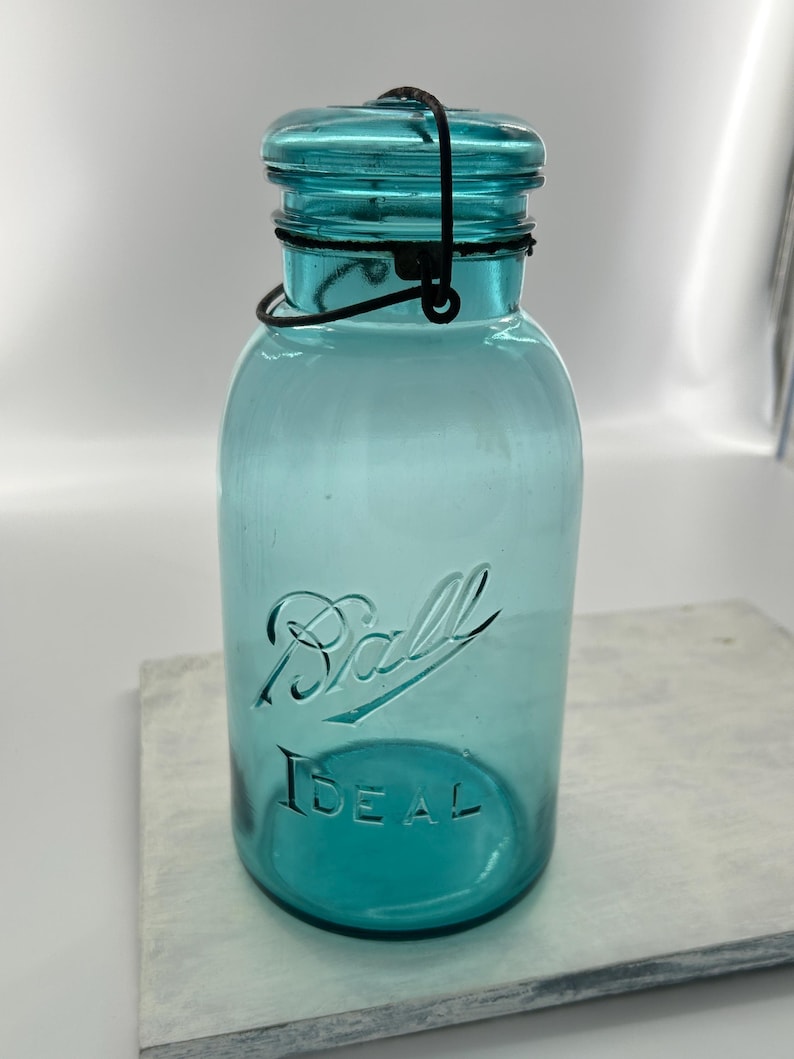 May include: A vintage teal-colored Ball Ideal jar with a matching lid and a black wire closure. The jar has the Ball Ideal logo embossed on the front. The jar is sitting on a light-colored surface.