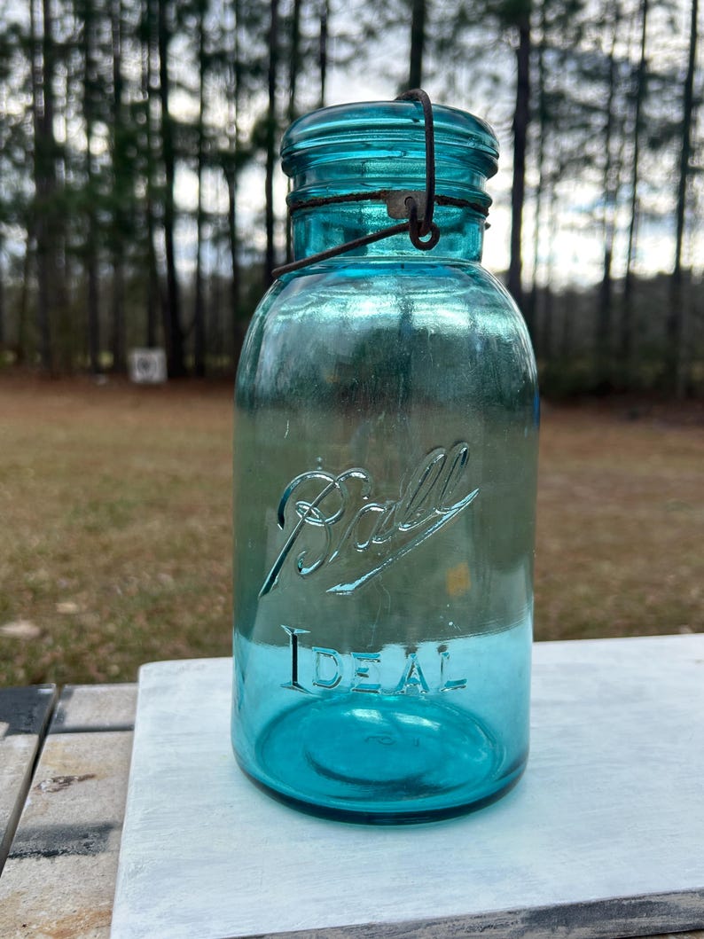 May include: A vintage, turquoise-colored Ball Ideal jar with a wire closure. The jar has the word "Ball" in script and "Ideal" printed on the front. The jar is made of glass and is sitting on a white surface.