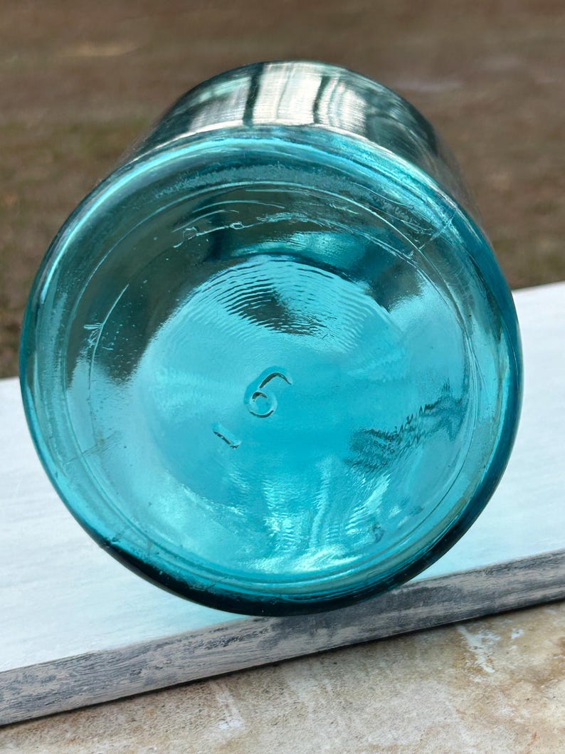 May include: A vintage, turquoise-colored glass jar, viewed from the bottom. The jar has a rounded base with the number "6" embossed. The glass is thick and has a slight texture. The jar is likely an antique or collectible item.