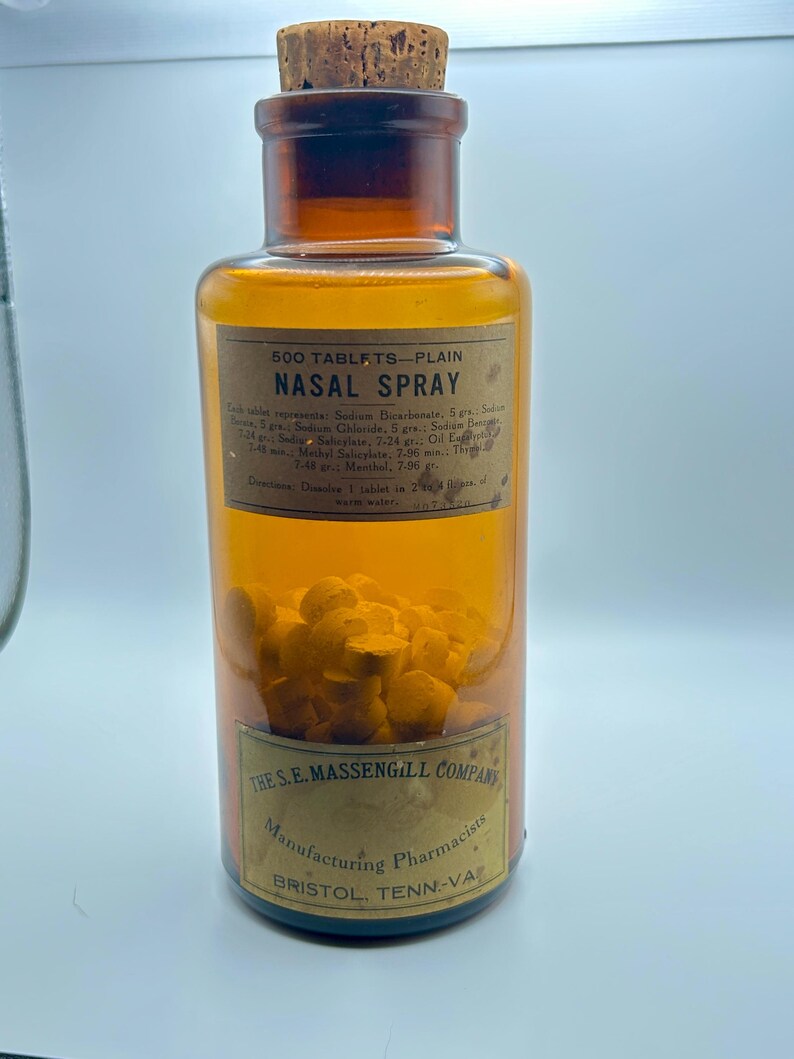 May include: An amber glass bottle with a cork stopper, labeled "NASAL SPRAY." The label includes text about ingredients and directions. The bottle contains numerous yellow tablets. The label also reads "THE S.E. MASSENGILL COMPANY, Manufacturing Pharmacists, BRISTOL, TENN-VA."