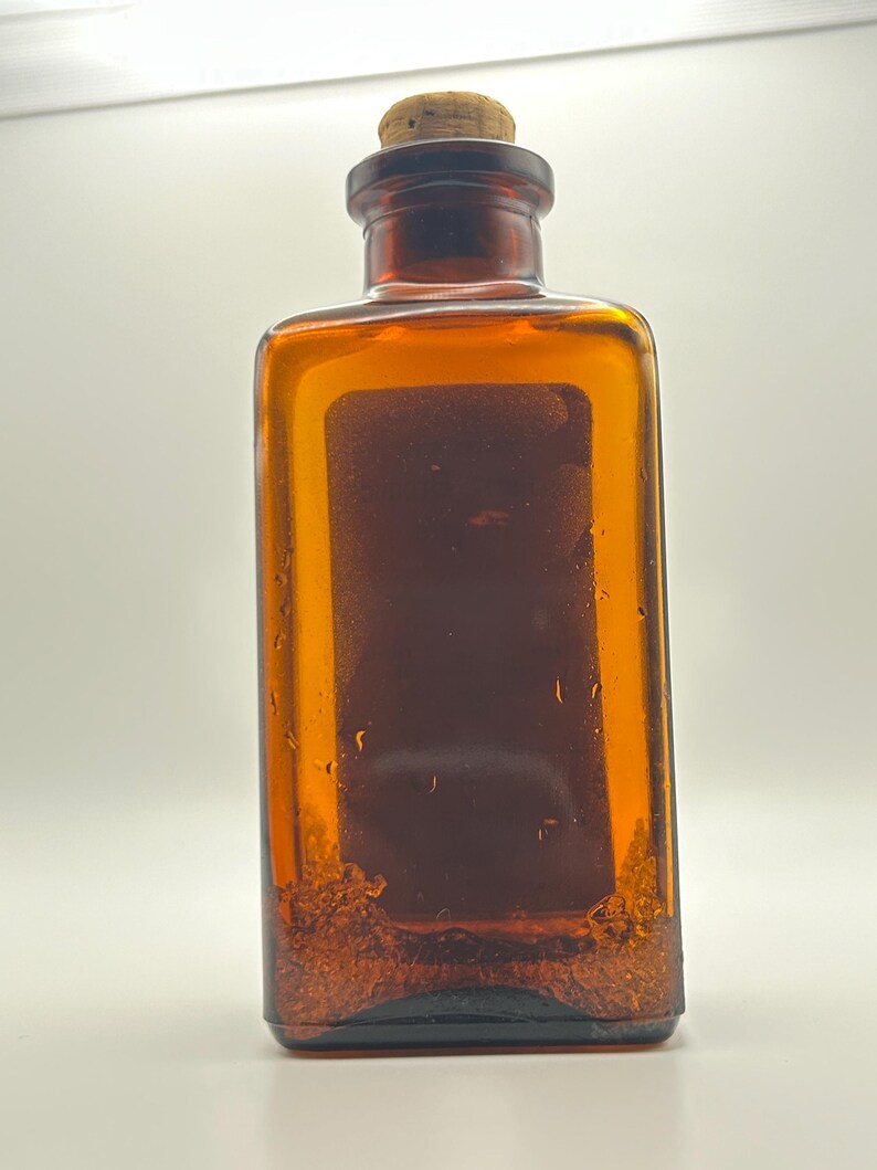 May include: Amber-colored rectangular glass bottle with a cork stopper. The bottle has a dark brown interior and appears to have sediment at the bottom. The bottle is likely vintage and could be used for decorative purposes.