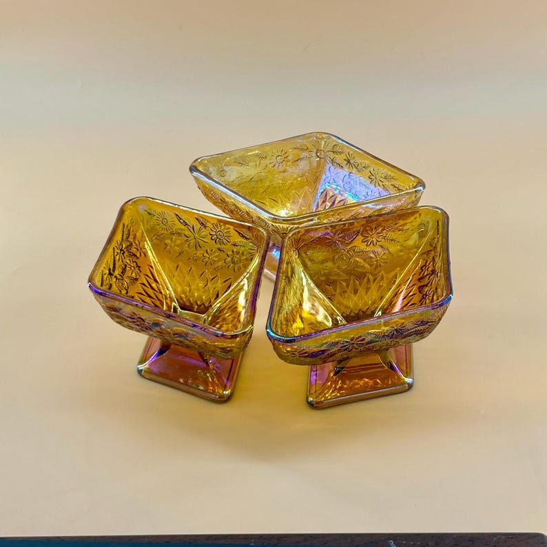 May include: Three iridescent amber-colored glass dessert dishes. Each dish has a square bowl shape with a floral pattern and sits on a square pedestal base. The glass reflects a rainbow of colors.