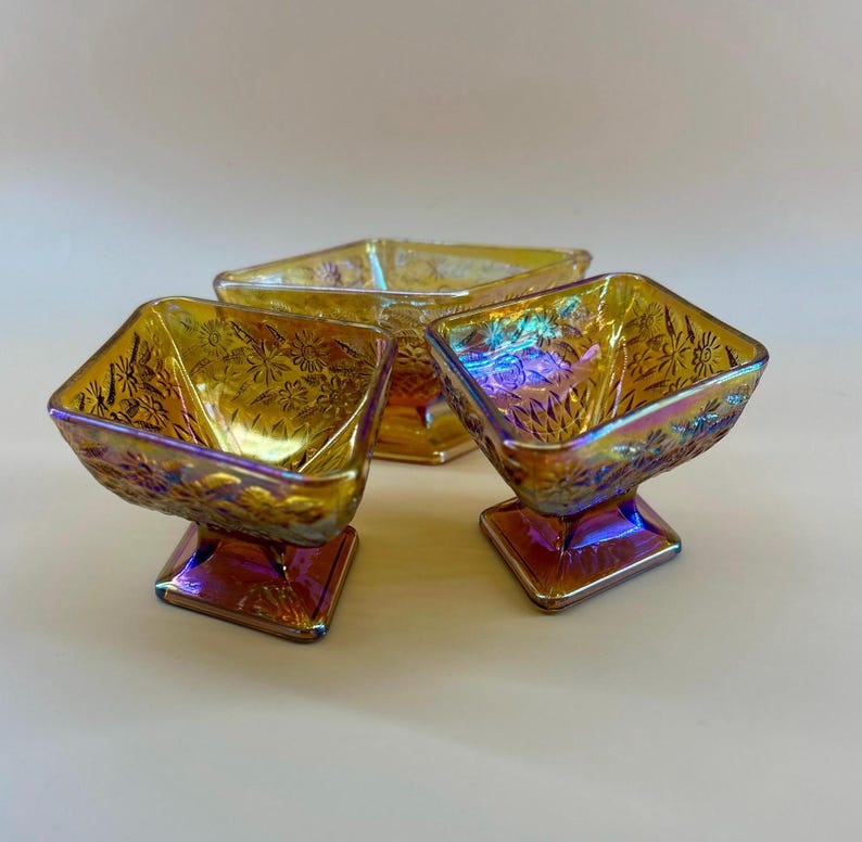 May include: Three iridescent glass dessert bowls. Each bowl has a square shape with a floral pattern and sits on a square pedestal base. The bowls have a golden-yellow color with hints of purple and blue.