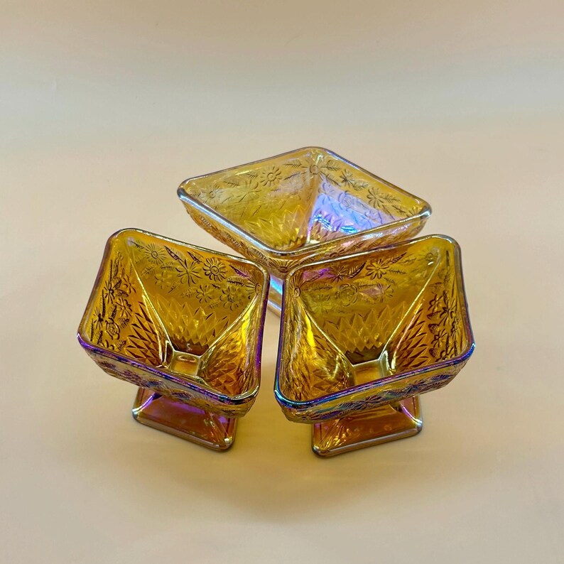 May include: Three iridescent amber glass dessert dishes. Each dish has a square bowl shape with a pedestal base. The glass has a floral pattern and a rainbow sheen. The dishes are arranged in a triangular formation.