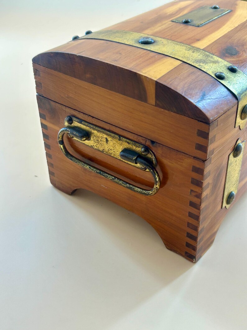 May include: A small, wooden chest with a curved top and brass hardware. The chest is made of light and dark brown wood, with a brass handle and metal bands. The chest has a rectangular shape and is likely used for storage.