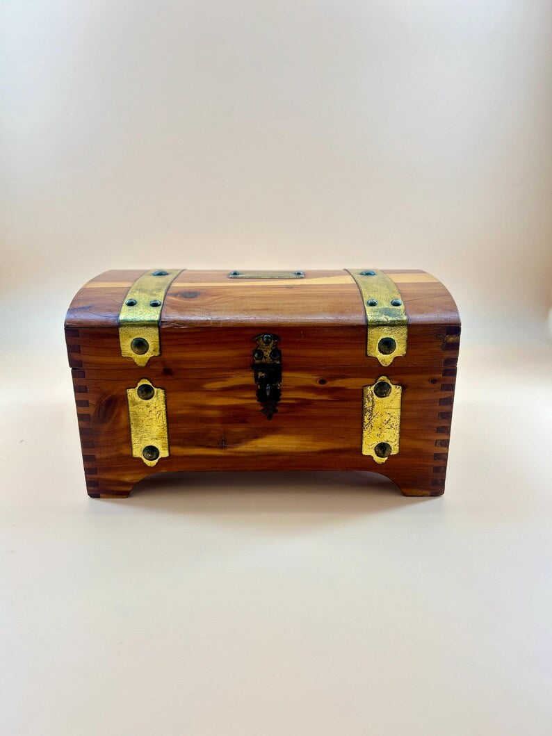 May include: A small, wooden chest with a curved top. The chest is made of reddish-brown wood and features brass-colored metal accents, including hinges, clasps, and decorative plates. The chest has a small, black metal latch and is elevated on short feet.