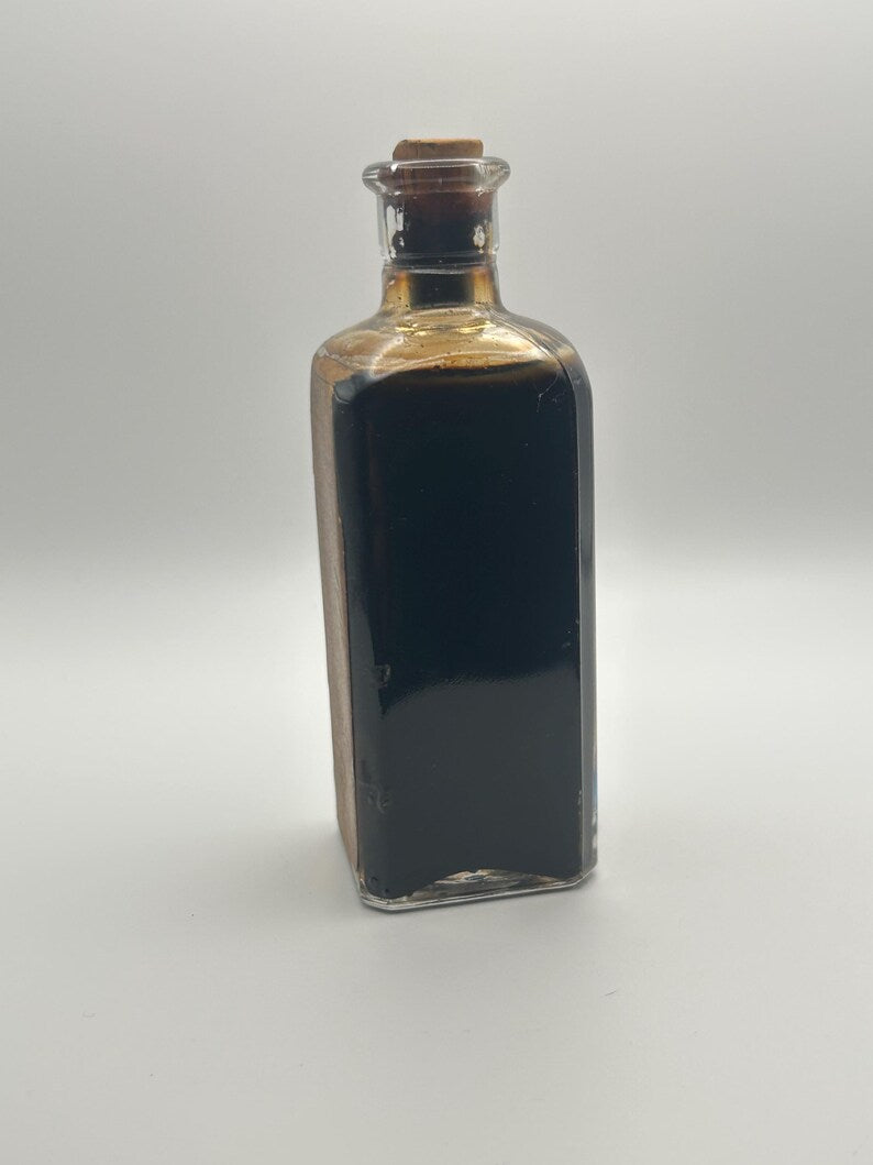 May include: A rectangular clear glass bottle filled with a dark brown liquid. The bottle features a narrow neck topped with a light brown cylindrical cork stopper. The glass container has distinct vertical edges and a flat base, holding dark contents.