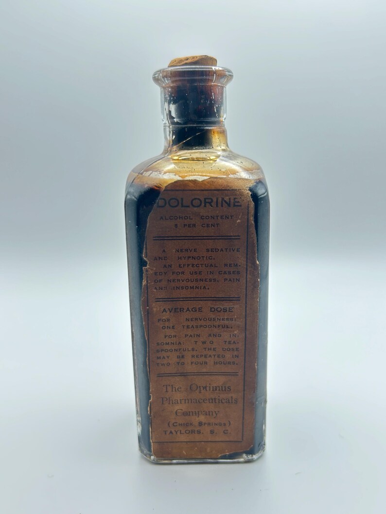 May include: A vintage rectangular glass bottle filled with a dark liquid, topped with a cork stopper. A tan paper label reads DOLORINE, ALCOHOL CONTENT 8 PER CENT, A NERVE SEDATIVE AND HYPNOTIC, AVERAGE DOSE, and The Optimus Pharmaceuticals Company.