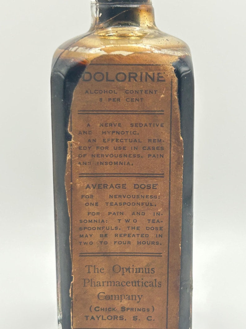 May include: A vintage rectangular glass bottle filled with a dark liquid, featuring a tan paper label with black text reading DOLORINE, ALCOHOL CONTENT 5 PER CENT, A NERVE SEDATIVE AND HYPNOTIC, AVERAGE DOSE, and The Optimus Pharmaceuticals Company.