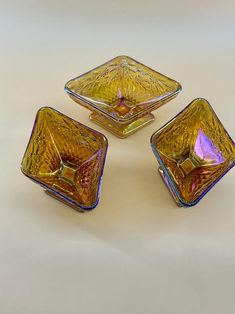 May include: Three iridescent glass serving dishes. Each dish is square with a diamond-shaped bowl and a pedestal base. The glass has a golden hue with hints of purple and blue. The dishes are decorated with a floral pattern.