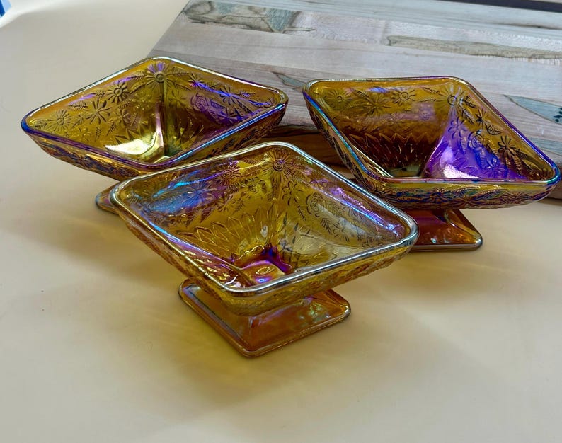 May include: Three iridescent glass serving dishes. Each dish has a square shape with a floral pattern and sits on a square pedestal base. The glass has a golden hue with hints of purple and blue.