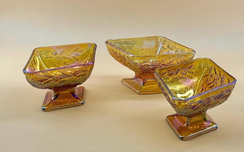 May include: Three iridescent glass dessert bowls with square bases and floral patterns. The bowls have a golden-yellow hue with hints of purple and orange. The bowls are elevated on a square pedestal base.