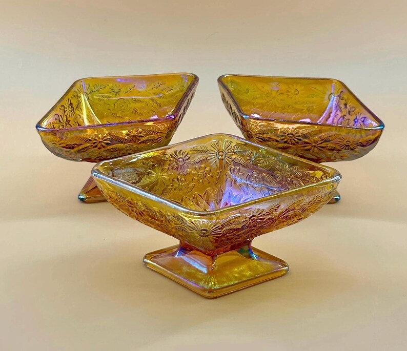 May include: Three iridescent amber glass dessert bowls with a square shape and floral pattern. The bowls are raised on a square base. The glass has a shiny, reflective surface.
