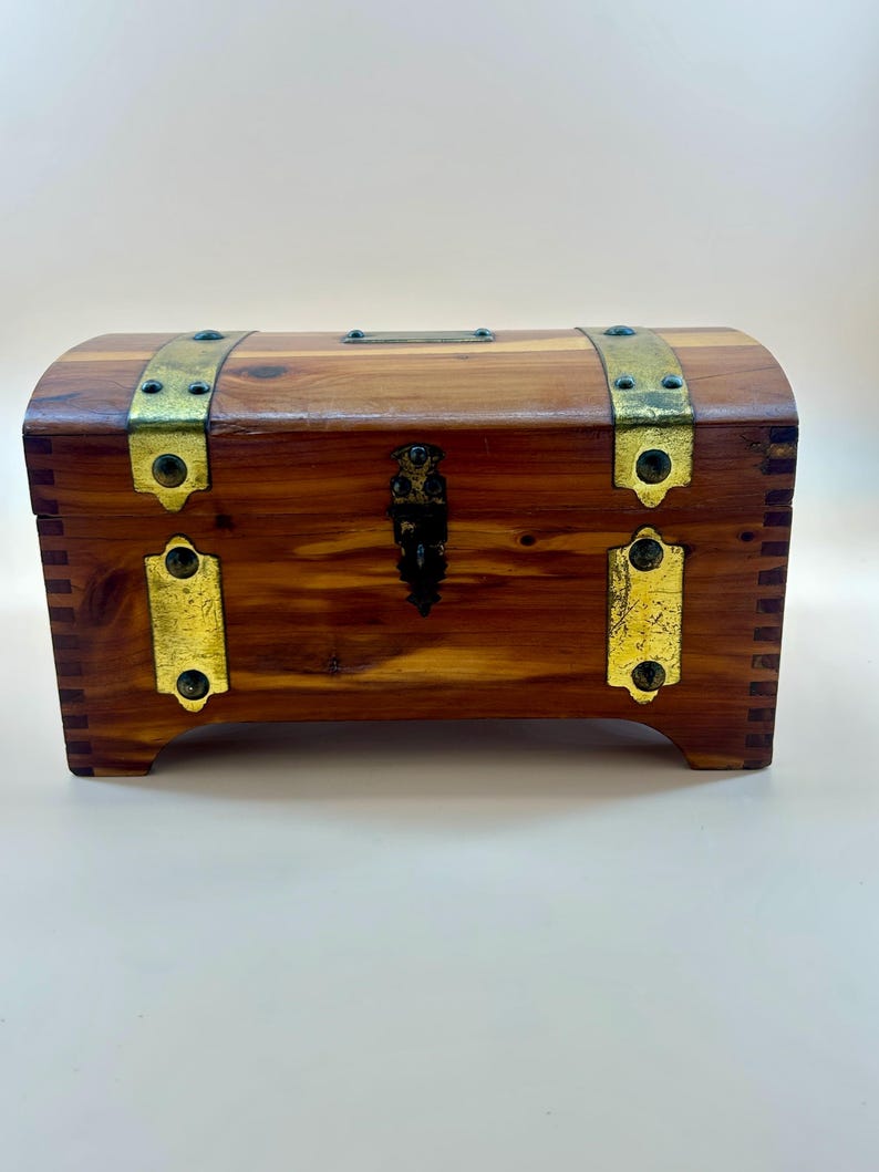 May include: A small, wooden chest with a curved top. The chest is made of reddish-brown wood and features brass-colored metal accents, including hinges, clasps, and decorative bands. The chest has a small, ornate lock and is raised on short feet.
