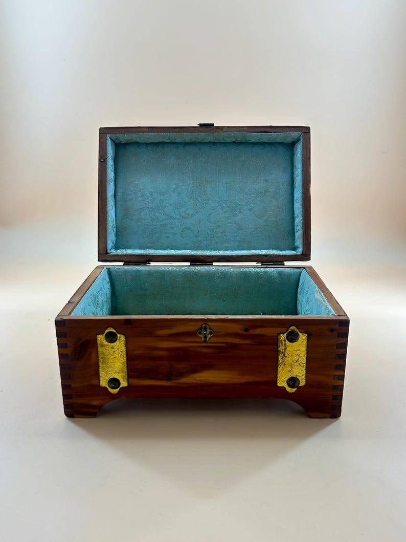 May include: Open wooden box with a hinged lid. The box is brown with gold-colored metal accents. The interior is lined with blue fabric with a subtle pattern. The box is rectangular and sits on small feet.