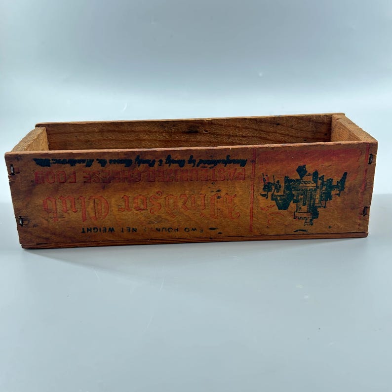 May include: A rectangular wooden crate with visible wear and tear. The crate has a faded red and green label with text that reads "Almsted's Club" and "Net Weight Two Pounds". The crate is empty and has a rustic, vintage appearance.
