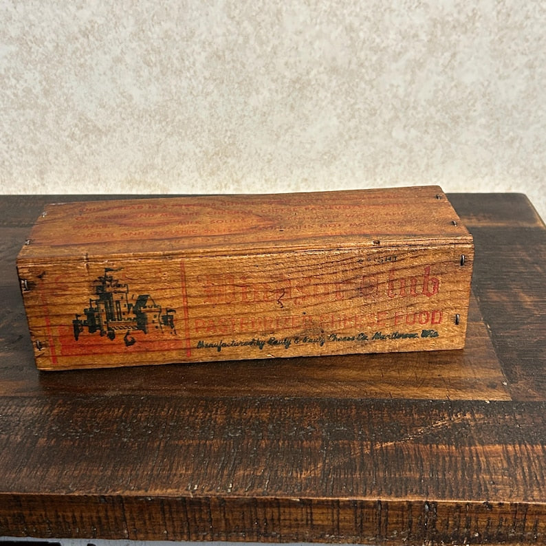 May include: A rectangular wooden box with a weathered appearance. The box has a red and black graphic on the front, along with text that reads "The Little Lamb Pasteurized Food". The box is sitting on a dark wooden surface.