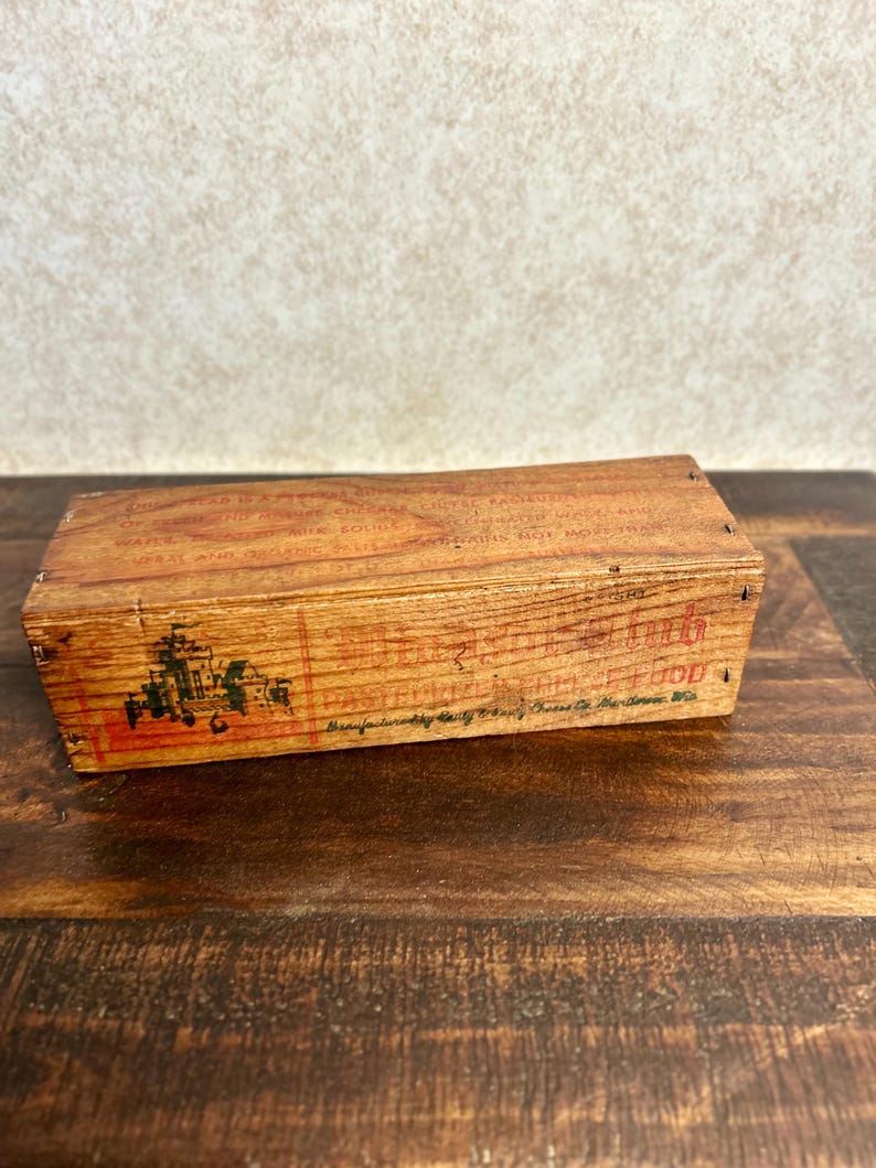 May include: A rectangular wooden box with faded red and black text and graphics. The box has a rustic, aged appearance, with visible wood grain and small nails. The text is partially legible, suggesting it may be a vintage product container.