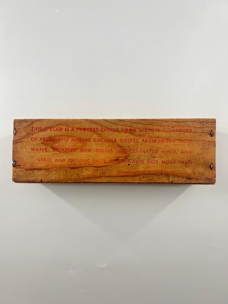 May include: A rectangular wooden box with red text. The text reads: "THIS SPREAD IS A PROCESS CHEESE FOOD MIXTURE COMPOSED OF FRESH AND MATURE CHEDDAR CHEESE PASTEURIZED WITH WATER..." The box is brown and has small nails.